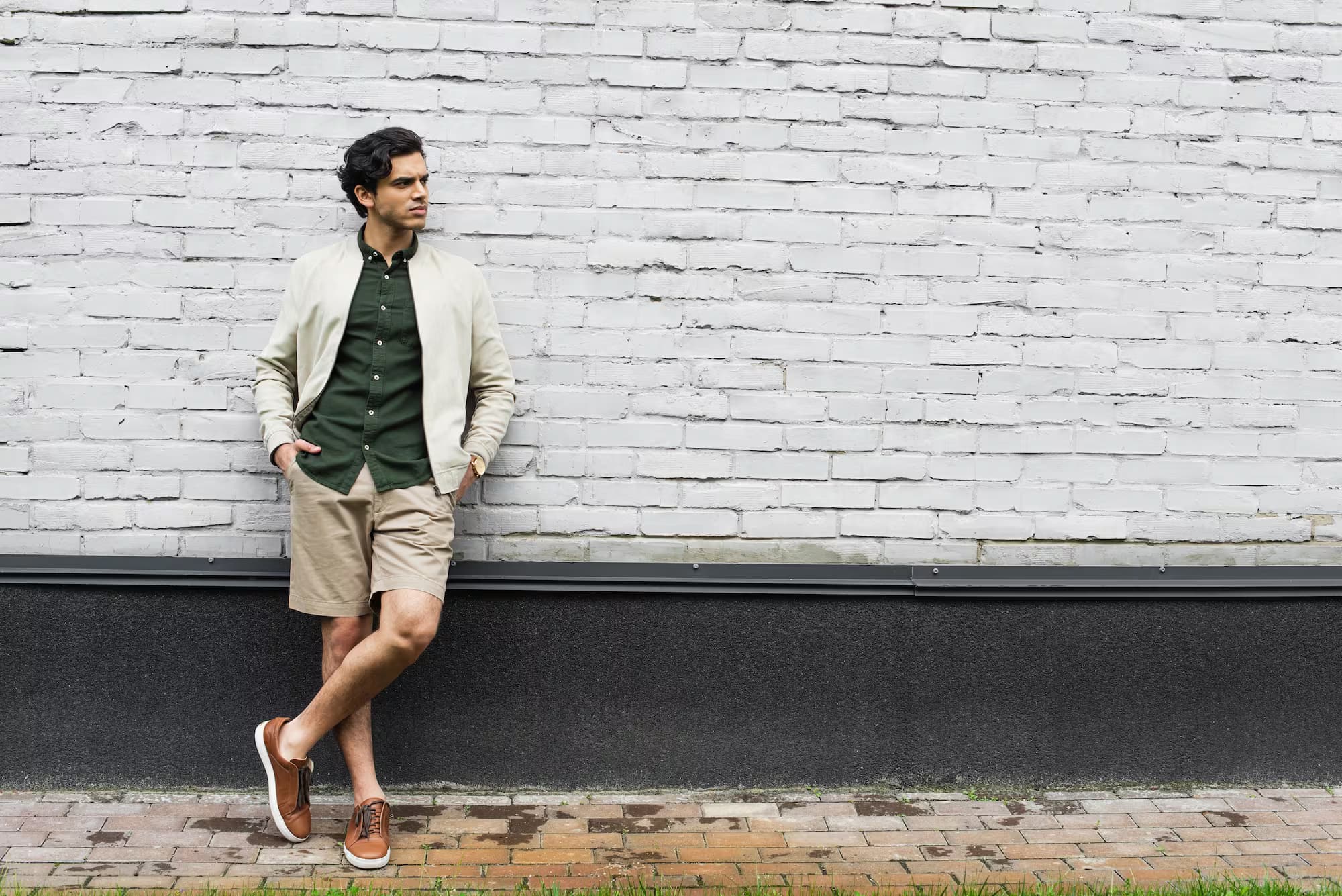 Man in a bomber jacket and shorts leaning against a white brick wall