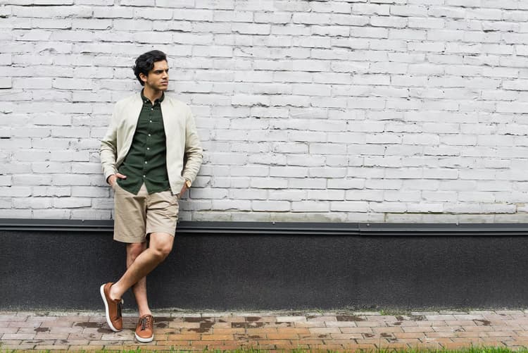 Man in a bomber jacket and shorts leaning against a white brick wall