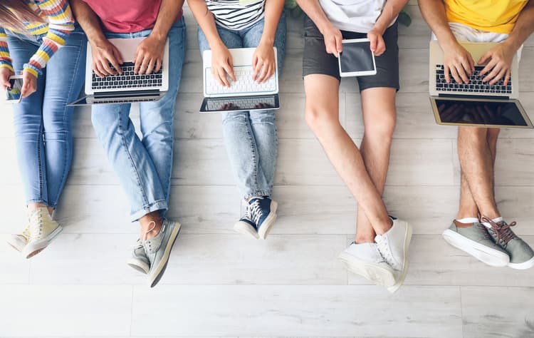Overhead view of four students seated in a row using laptops, a tablet, and a phone